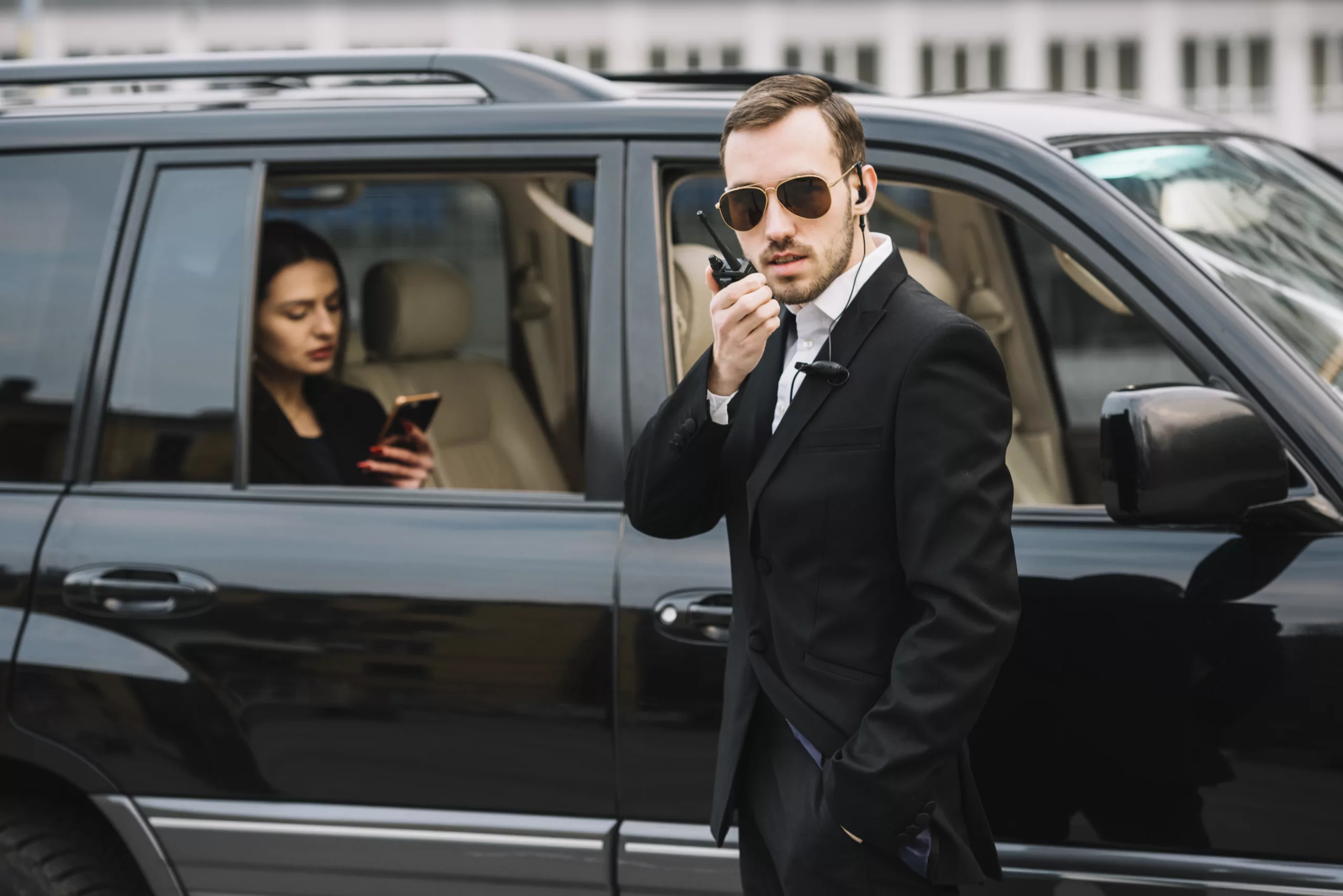 Security professional in a suit communicates via earpiece next to a luxury black SUV, providing vip security services by HK Defense Solutions.