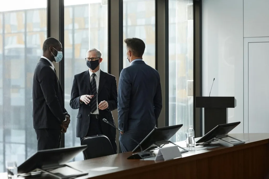 Three professionally dressed men wearing face masks and suits have a discussion in a conference room with large glass windows, accompanied by computer monitors and a podium, reflecting a secure corporate environment.